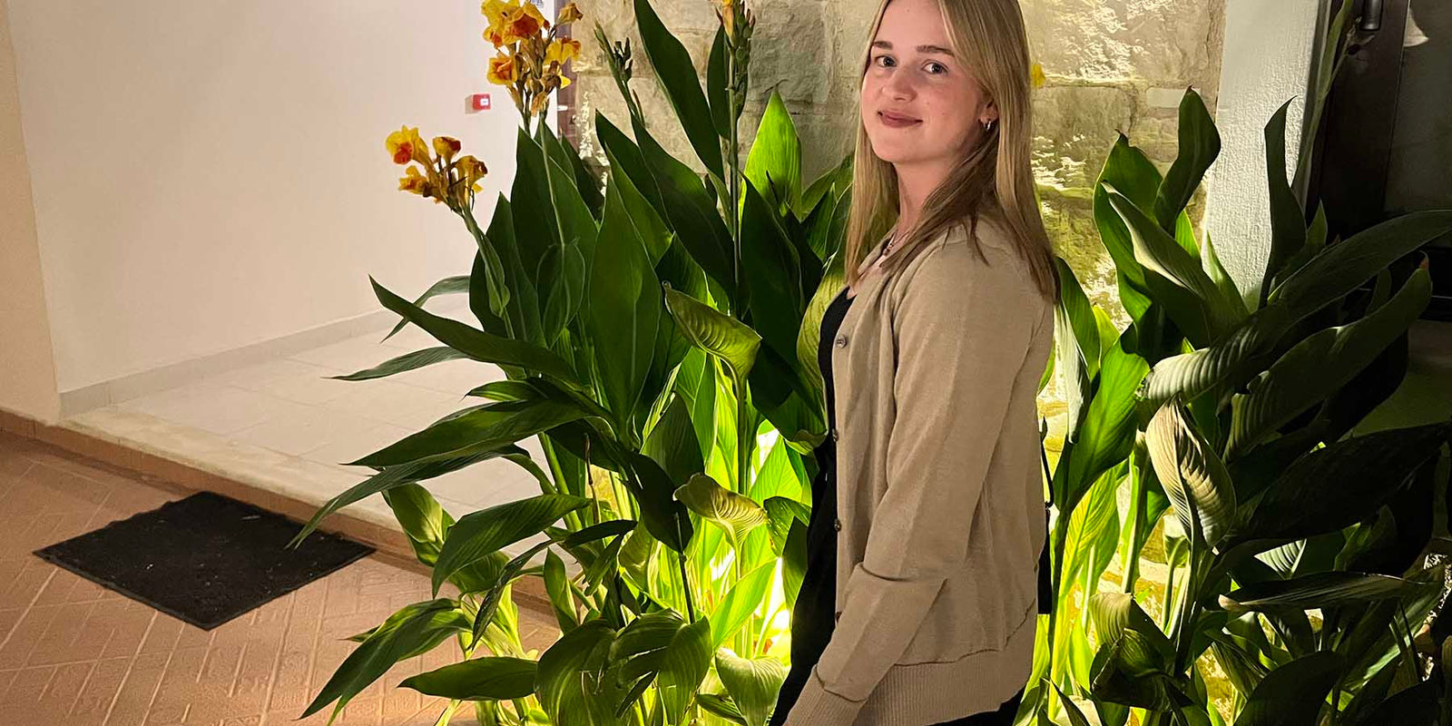 Alpakka neuletakki hiekka Trujillo nuoren naisen päällä, taustalla viherkasveja.